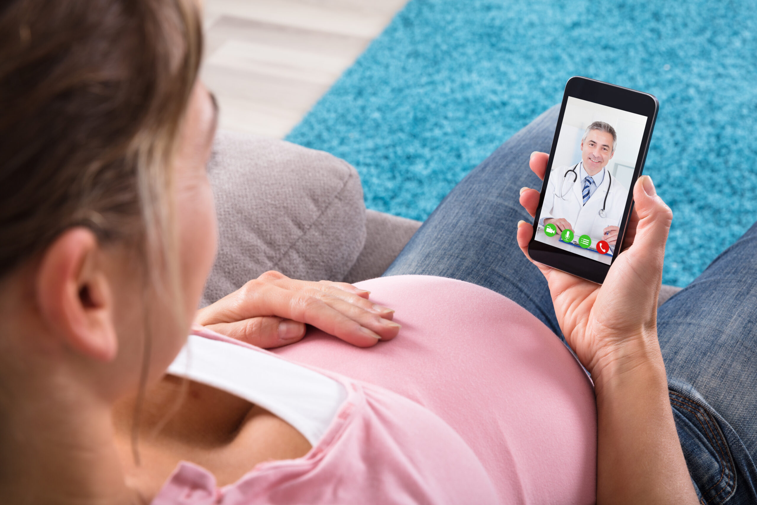 Expecting Pregnant Woman Sitting On Sofa Video Conferencing With Doctor On Mobile Phone